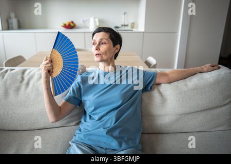 Exhausted mature woman fanning herself at home. Unbearable heat, elderly suffering from stuffy Stock Photo
