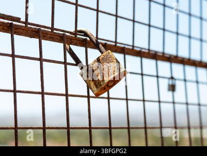 A rusty padlock on a weathered metal fence—likely placed as a sentimental symbol. In the background, another padlock hangs slightly out of focus. Stock Photo