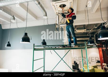 Worker performs maintenance on HVAC system while standing on scaffolding in an office space during the daytime Stock Photo