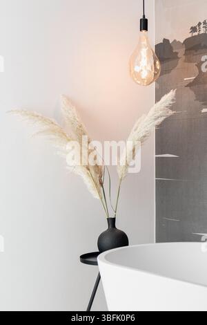 Modern bathroom featuring a white bathtub, pampas grass in a black vase, and a stylish hanging light bulb Stock Photo