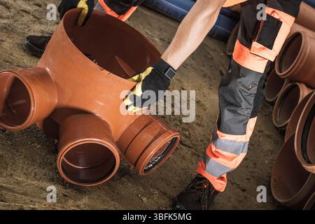 A construction worker handles a large drainage pipe fitting with precision while surrounded by additional pipes. The site is bustling with activity an Stock Photo