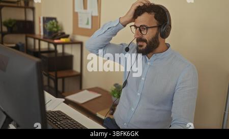Hispanic man with beard wearing headset in office setting, engaging through computer for virtual communication, emphasizing modern workspace atmospher Stock Photo