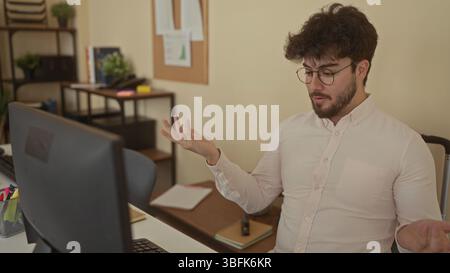 Young man with beard working at computer in office room, looking thoughtful and engaged, surrounded by books and plants, creating a casual yet profess Stock Photo