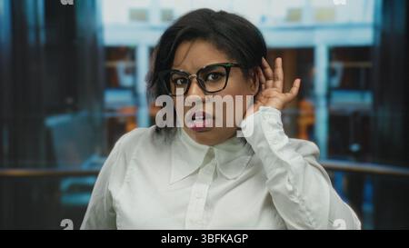 Woman wearing glasses in hotel indoors listening intently with hand on ear suggesting curiosity professional setting Stock Photo
