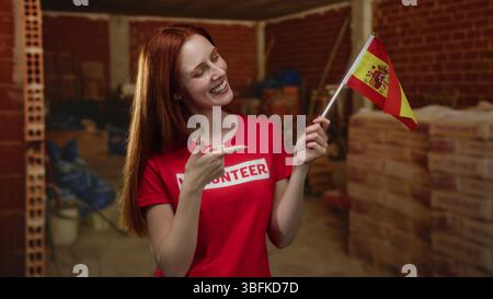 Woman smiling holding spain flag at indoor construction site highlighting volunteer work with vibrant red shirt and joyful expression in a rustic back Stock Photo