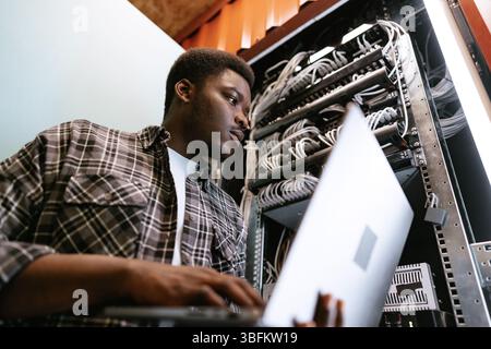 IT professional manages server rack while working on laptop in a data center during afternoon hours Stock Photo