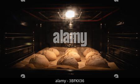 Rustic sourdough rolls rising on a baking tray under warm oven light, seen through the oven door with visible heater elements. Kitchen scene. Stock Photo