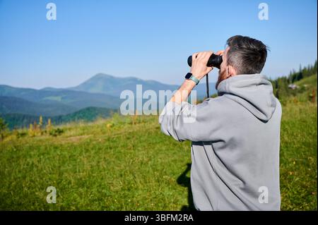 Man stands in grassy field, using binoculars to observe the distant mountains, wearing gray hoodie, smartwatch on his left wrist. Rolling green hills and clear blue sky, creating tranquil scene. Stock Photo