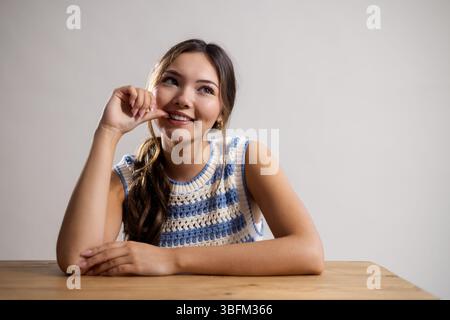 Waist-up studio portrait of a young adult female sitting at a table, looking up to one side and smiling. She is leaning both arms on the table, one th Stock Photo