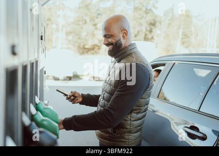 Smiling mature man with shaved head making online payment while standing near car at gas station Stock Photo