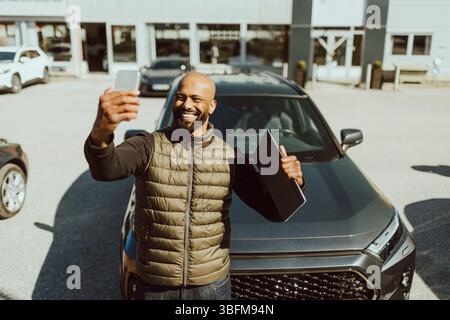 Happy mature man taking selfie through smart phone with new car outside showroom Stock Photo
