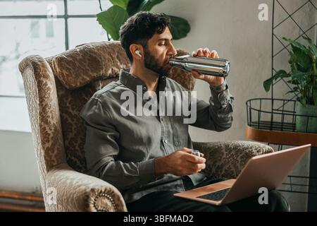 Thirsty businessman drinking water while sitting on armchair with laptop at office Stock Photo