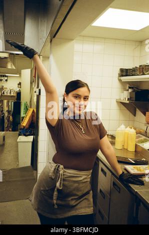 Portrait of smiling young woman standing with hand raised in restaurant kitchen Stock Photo