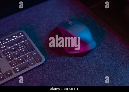 A detailed close-up of an ergonomic computer mouse and a portion of a backlit keyboard, bathed in vibrant pink and blue ambient lighting on a dark des Stock Photo