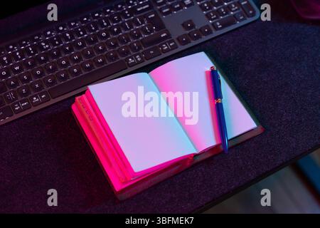 An open notebook with blank pages and a pen, alongside a computer keyboard, illuminated by vibrant pink and blue ambient lighting on a dark desk. Idea Stock Photo