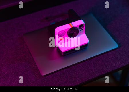 A retro-style instant camera placed on top of a closed laptop, bathed in vibrant pink and purple ambient lighting on a dark desk. Symbolizes a blend o Stock Photo