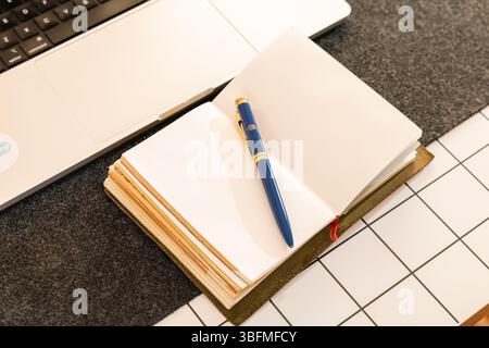 Top-down view of an open notebook with blank pages and a classic blue and gold pen, placed beside a laptop on a desk with a felt mat and gridded surfa Stock Photo