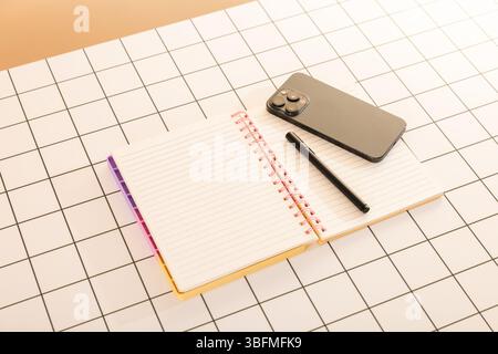 Top-down flat lay of an open spiral notebook with lined paper, a black pen, and a smartphone, arranged on a white surface with a black grid pattern. T Stock Photo