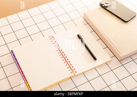 Organized flat lay of an open spiral notebook, a closed journal or book, a black pen, and a smartphone on a white and black gridded desktop. Represent Stock Photo