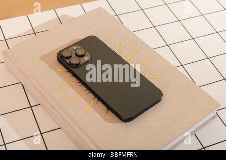 Close-up flat lay of a contemporary dark smartphone resting on a closed book or journal featuring a golden sunburst design, set against a white and bl Stock Photo