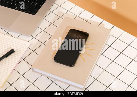Top-view of an organized desk with a smartphone displaying the time on a book with a sunburst design, alongside a laptop and an open notebook with a p Stock Photo