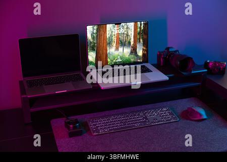 Wide shot of a modern creator's desk featuring a dual laptop setup on a riser, professional and vintage cameras, keyboard, and mouse, all under immers Stock Photo