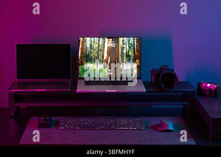 Full view of a sophisticated battlestation or creator workspace, dramatically lit by intense blue and pink neon lights. Features dual laptops on a ris Stock Photo