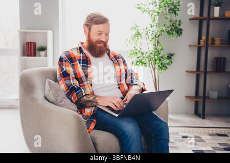Middle aged man with a beard smiling while working on a laptop, comfortably seated in a modern living room during daylight. Stock Photo