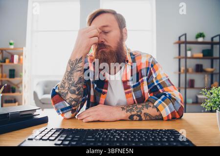 Bearded man wearing a checkered shirt sitting at a wooden desk indoors expressing stress or contemplation while in daylight Stock Photo
