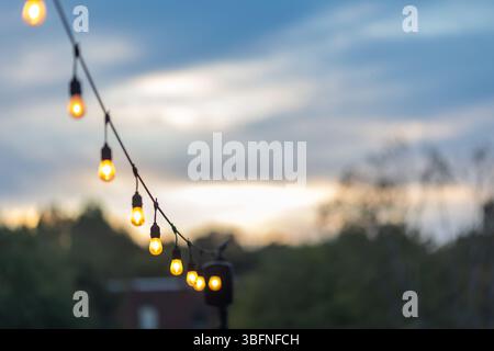 A string of glowing edison light bulbs hung on a patio creates a festive outdoor vibe against a twilight sky. Stock Photo