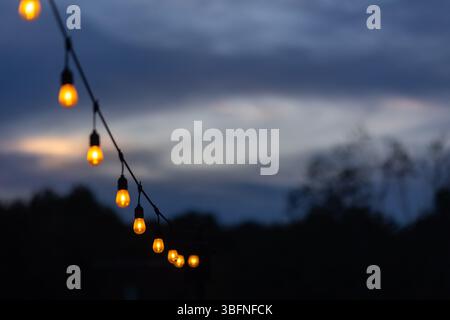 A string of glowing edison light bulbs hung on a patio creates a festive outdoor party vibe against a purple dusk sky. Stock Photo