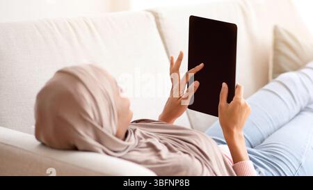 African American woman in hijab with tablet computer lying on sofa at home, mockup for design on screen Stock Photo
