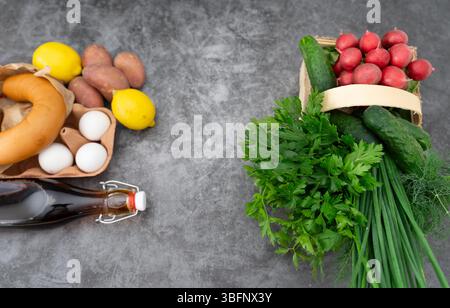 A bottle of kvass sits among fresh okroshka ingredients on a rich gray wooden table. Stock Photo