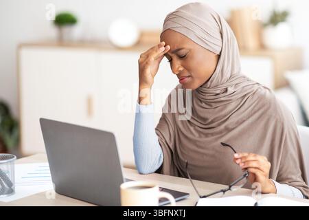 Deadline stress. Exhausted black woman in hijab tired after working on laptop for too long, having headache at home Stock Photo