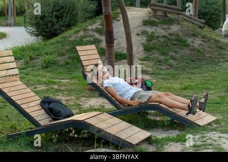 Relaxed boy in glasses lying on wooden bench in park on summer day.  Stock Photo