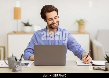 Cheerful Businessman Working At Laptop Taking Notes Sitting At Workplace Stock Photo