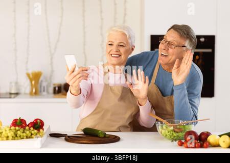 Senior Couple Making Video Call Using Mobile Phone In Kitchen Stock Photo