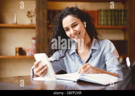 Woman sitting at desk, using smartphone and writing in notebook Stock Photo