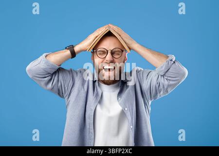 Furious young man putting book on his head and screaming on blue studio background Stock Photo