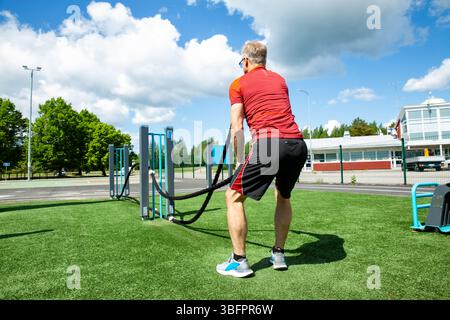 Active middle-aged man training with battle ropes at an outdoor gym on a sunny summer day. Fitness, strength, and healthy lifestyle in the open air Stock Photo