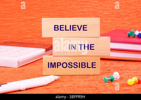 Motivational quote. BELIEVE IN THE IMPOSSIBLE text made of wooden blocks on an orange background with stationery lying next to it. Stock Photo