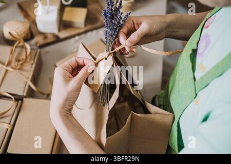 Hands tie kraft gift bag with lavender and ribbon in a natural packaging setup. Eco-friendly branding, sustainable wrapping, plastic-free, zero-waste Stock Photo
