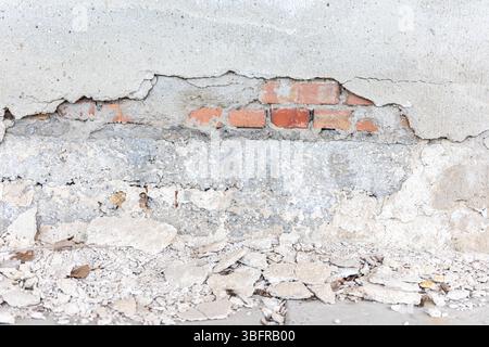 Close-up of a deteriorating concrete wall with crumbling plaster revealing red bricks and scattered debris on the ground. Stock Photo