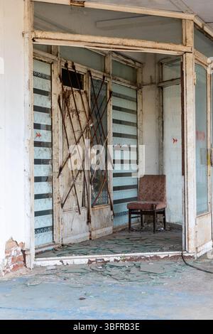 Dilapidated shop entrance with shattered glass, rusted metal bars, and a worn-out chair, evoking decay and abandonment. Stock Photo