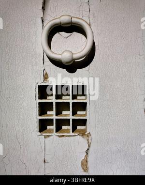 A striking white door knocker adorns a weathered wall featuring small square openings arranged in an organized grid. Stock Photo