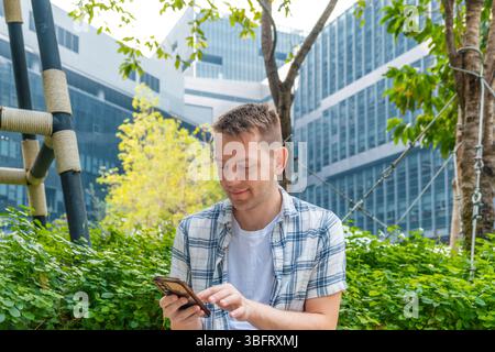 Focused young man sitting outdoors using smartphone in urban background with greenery. Male using mobile phone in green city. Modern city life Stock Photo