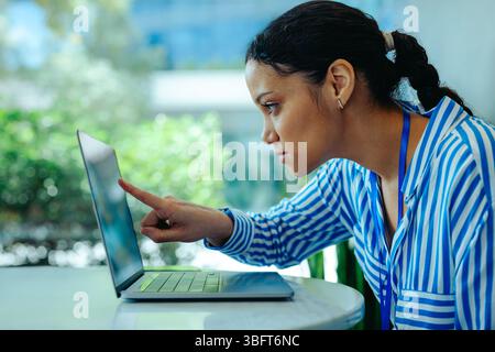 A focused corporate accountant in a striped blue shirt intensely analyzes data on her laptop, pointing at the screen. Concentration and professionalis Stock Photo