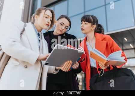 Three businesswomen collaborating outdoors while reviewing notes and using digital devices Stock Photo