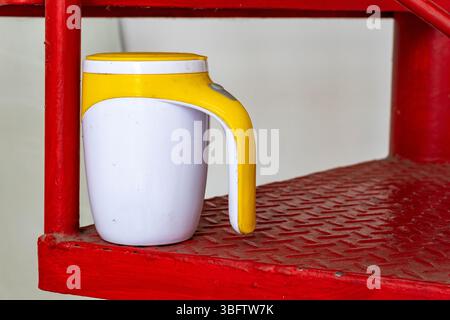 White and yellow coffee cup sits on a red surface. The cup is placed on a stairway, and the red surface is made of metal. Concept of warmth and comfor Stock Photo