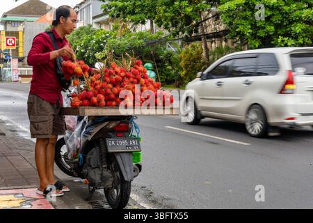 Bali, Indonesia - Dec 10, 2024: Man is selling fruit called rambutan on a motorcycle. The fruit is red and there are many of them Stock Photo